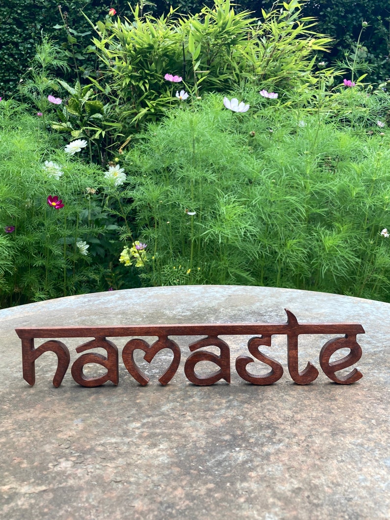 May include: A wooden sign with the word "namaste" in a stylized font. The sign is a dark brown color and sits on a stone surface, with a lush green garden and flowers in the background.