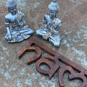 May include: Two silver-colored Buddha statues in a seated position with hands in prayer. The statues have intricate details and are adorned with sparkling embellishments. Below the statues are wooden cut-out letters in a dark brown color.