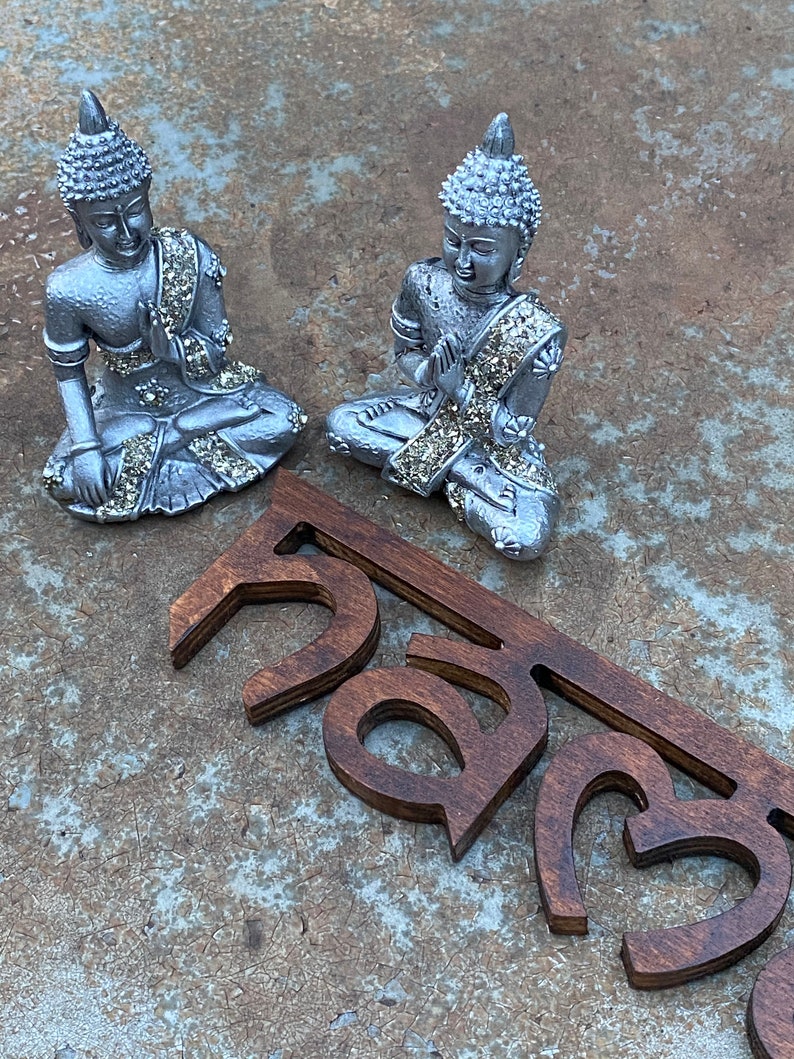 May include: Two silver-colored Buddha statues in a seated position with hands in prayer. The statues have intricate details and are adorned with sparkling embellishments. Below the statues are wooden cut-out letters in a dark brown color.