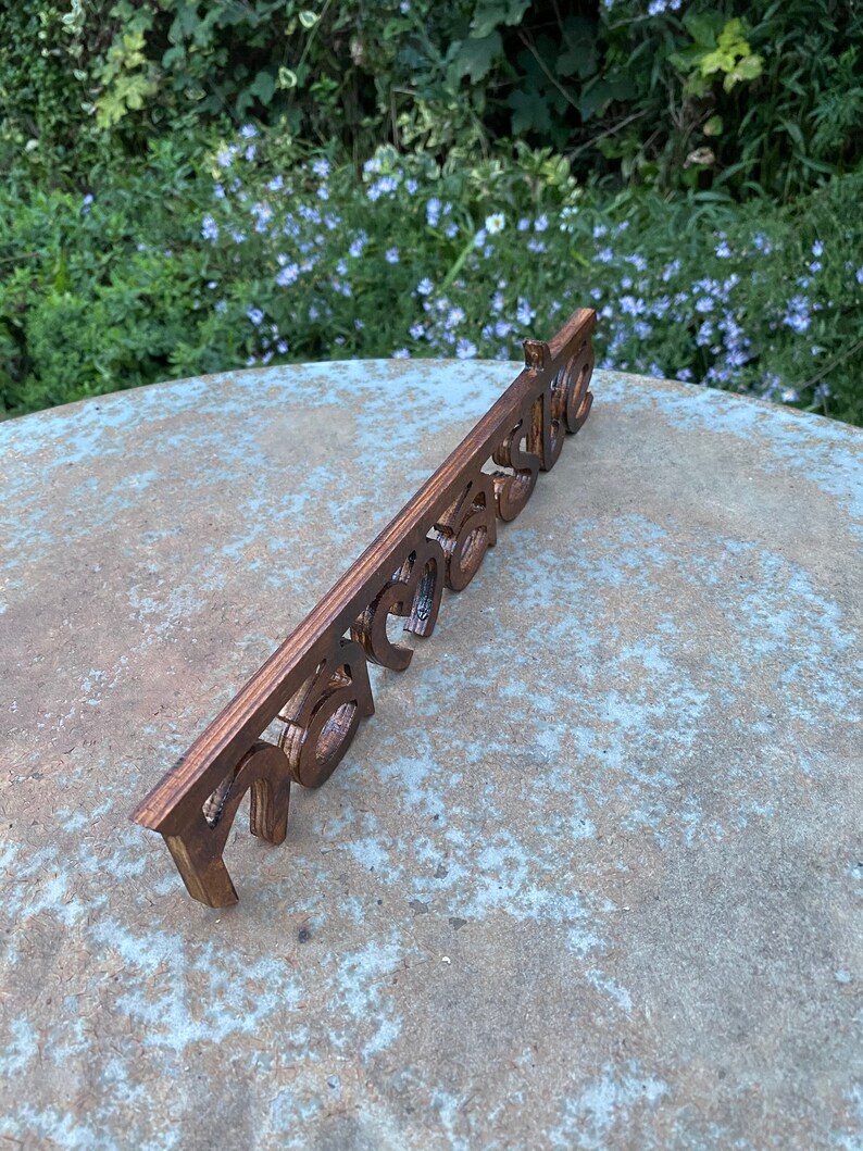 May include: A wooden sign with the word "Welcome" cut out in a decorative font. The sign is a warm brown color and is placed on a stone surface with a natural background of green foliage and blue flowers.