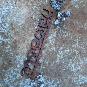 May include: A wooden sign with the word "Namaste" cut out in a dark brown color, accompanied by three small silver Buddha statues. The statues are in a seated meditation pose. The background is a textured, mottled gray surface.
