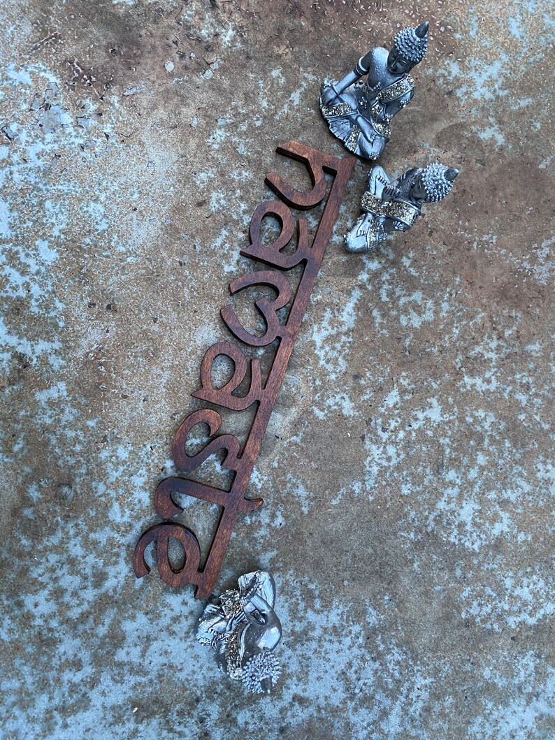 May include: A wooden sign with the word "Namaste" cut out in a dark brown color, accompanied by three small silver Buddha statues. The statues are in a seated meditation pose. The background is a textured, mottled gray surface.
