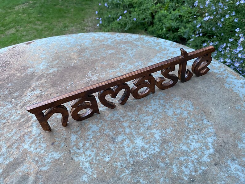 May include: A wooden sign with the word "Namaste" in a stylized font. The sign is made of dark brown wood and is placed on a light gray surface. The background includes green grass and foliage.