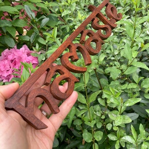 May include: A wooden sign in a dark brown stain, featuring the word "Raise" in a stylized font. The sign is held against a backdrop of green foliage and pink flowers.