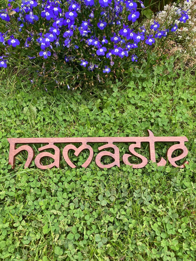 May include: A wooden sign with the word "namaste" in a stylized font. The sign is a warm brown color and is placed on a bed of green grass and clover, with a backdrop of vibrant blue flowers.