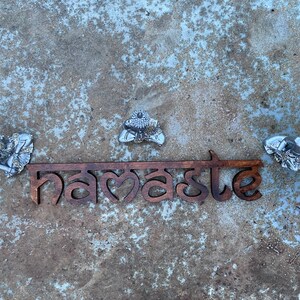 May include: A wooden sign with the word "namaste" in a stylized font, flanked by three silver Buddha figures. The sign is brown, and the Buddhas are detailed with intricate designs. The background is a textured, mottled gray surface.