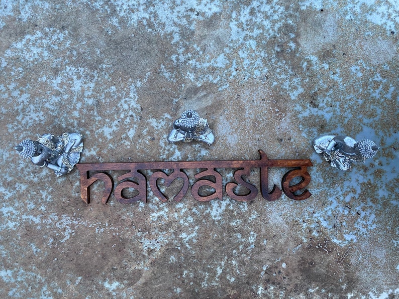 May include: A wooden sign with the word "namaste" in a stylized font, flanked by three silver Buddha figures. The sign is brown, and the Buddhas are detailed with intricate designs. The background is a textured, mottled gray surface.
