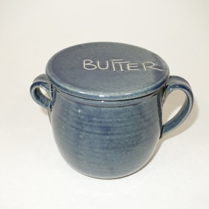 Könnte beinhalten: Eine blaue Keramik-Butterdose mit Deckel. Auf dem Deckel steht das Wort "BUTTER". Die Dose hat zwei Griffe und eine abgerundete Form. Die Butterdose hat eine matte blaue Farbe.