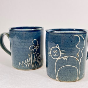 Könnte beinhalten: Zwei blaue Keramikbecher. Einer zeigt einen weißen Umriss einer Katze, der andere weiße Umrisse von Blumen und Gras. Beide Becher haben einen Henkel und ein leicht gesprenkeltes Aussehen.
