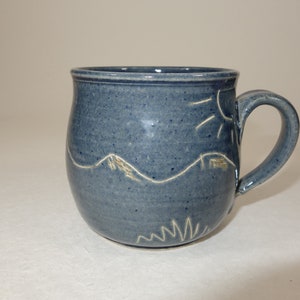 Könnte beinhalten: Eine blaue Keramiktasse mit einem weißen Sonnen-, Berg- und Grasdesign.