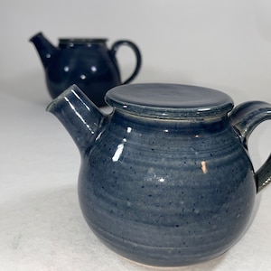 Könnte beinhalten: Eine blaue Teekanne aus Keramik mit Deckel und Henkel. Die Teekanne hat eine gesprenkelte Glasur und einen runden Korpus.