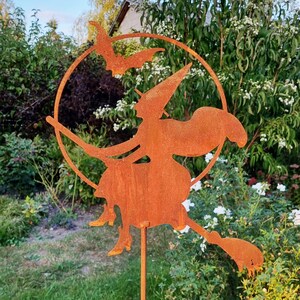 May include: A rusty metal silhouette of a witch flying on a broomstick with bats in the background. The witch is wearing a pointed hat and a long flowing robe. The silhouette is mounted on a metal stake.