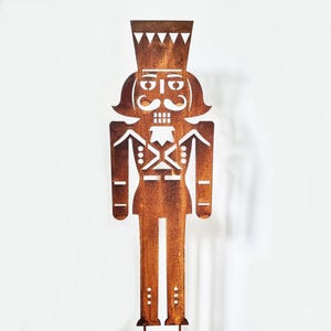 Könnte beinhalten: Eine rostige Nussknacker-Silhouette aus Metall mit einem weißen Design. Der Nussknacker steht hoch und hat eine Krone, einen Schnurrbart und eine detaillierte Uniform.