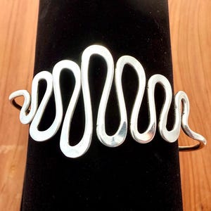 Può includere: Un bracciale a polsino color argento con un design aperto e fluido. Il bracciale presenta una serie di anelli ondulati interconnessi, creando un'estetica fluida e moderna. Il metallo ha una finitura lucida, che riflette la luce. Il bracciale è esposto su una superficie nera.