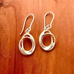 Pode incluir: Um par de brincos de prata polida com um design oval aberto e torcido. Cada brinco pende de um gancho de prata curvo, exibindo uma superfície metálica lisa e reflexiva com linhas fluidas e contínuas que formam um formato circular.