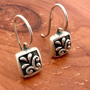 Peut inclure: Une paire de boucles d'oreilles pendantes en argent avec des pendentifs carrés aux coins arrondis. Chaque pendentif présente un motif floral en volutes d'argent poli en relief sur un fond encastré et sombre. Les boucles pendent à des crochets en argent.