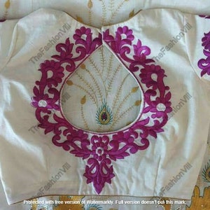 Op de afbeelding: Een wit blouse met een roze bloemenborduursel rond de halslijn. De borduurwerk heeft een groot, open, cirkelvormig ontwerp in het midden.