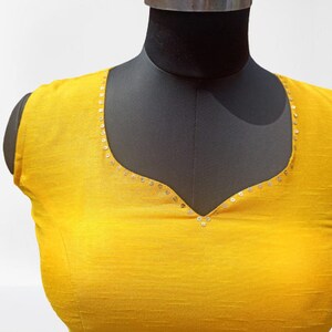 May include: A vibrant yellow blouse with a scoop neckline, presented on a black mannequin. The neckline is delicately adorned with small, silver sequins. The fabric has a subtle texture. The overall style is simple and elegant.