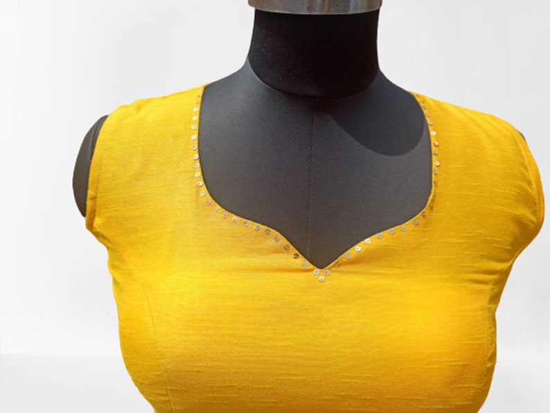 May include: A vibrant yellow blouse with a scoop neckline, presented on a black mannequin. The neckline is delicately adorned with small, silver sequins. The fabric has a subtle texture. The overall style is simple and elegant.