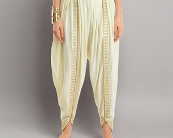 May include: Off-white dhoti trousers featuring a high-waisted design with vertical panels of gold-toned embroidery and small circular mirror accents running down the front of each leg, ending in a tapered hem with matching gold trim.