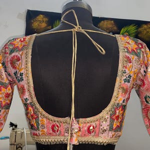 May include: A pink embroidered blouse with colorful floral and geometric patterns. The blouse has a low back with a tie closure. The sleeves are three-quarter length and are also embroidered.