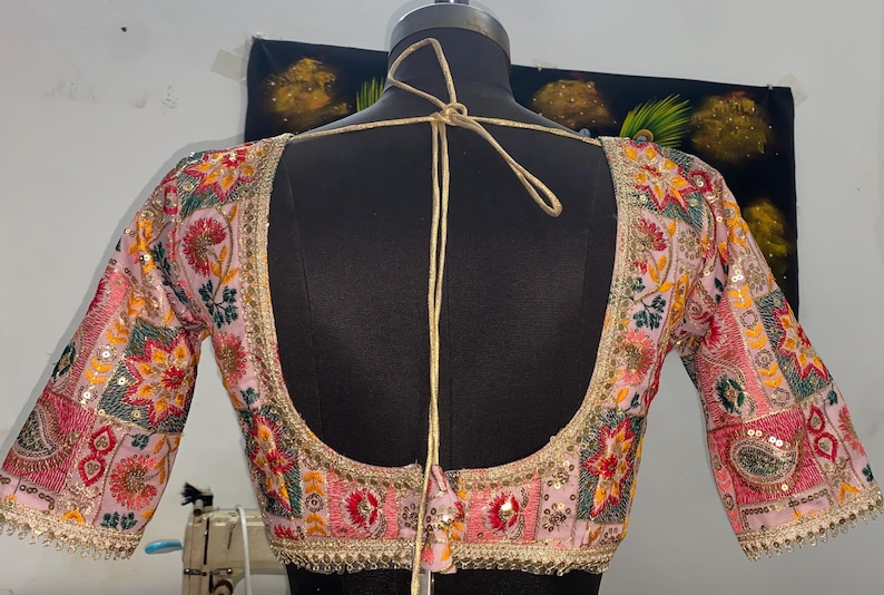 May include: A pink embroidered blouse with colorful floral and geometric patterns. The blouse has a low back with a tie closure. The sleeves are three-quarter length and are also embroidered.
