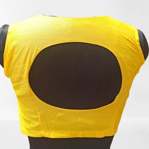 May include: A vibrant yellow blouse featuring a large oval cut-out at the back. The garment is displayed on a black mannequin, highlighting the back design. The fabric has a textured finish.