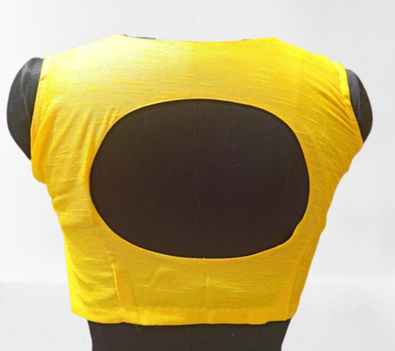 May include: A vibrant yellow blouse featuring a large oval cut-out at the back. The garment is displayed on a black mannequin, highlighting the back design. The fabric has a textured finish.