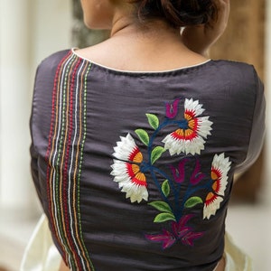 Pode incluir: Uma blusa cinza escura com um design de bordado floral branco, verde, laranja e roxo. A blusa tem uma bainha branca ao redor do decote e uma fileira de costuras coloridas nas costas.