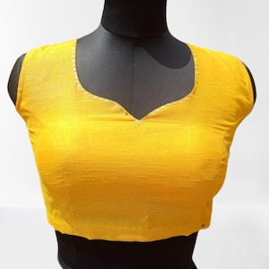 May include: A sleeveless, yellow blouse with a V-neck. The neckline is decorated with small, silver sequins. The blouse is displayed on a black mannequin against a white background. The fabric has a subtle texture.