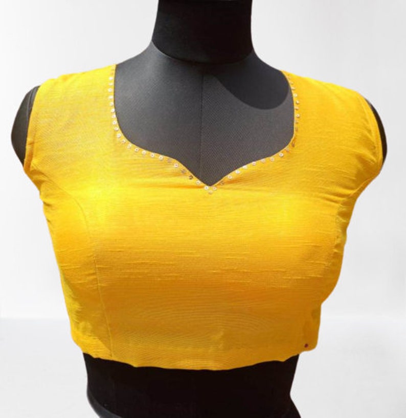 May include: A sleeveless, yellow blouse with a V-neck. The neckline is decorated with small, silver sequins. The blouse is displayed on a black mannequin against a white background. The fabric has a subtle texture.