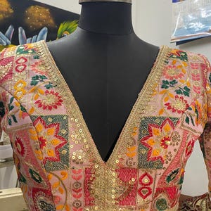 May include: A pink, embroidered blouse with a deep V-neckline. The blouse features a patchwork design with floral and geometric motifs in shades of red, green, yellow, and gold. The neckline and sleeves are trimmed with gold sequins.