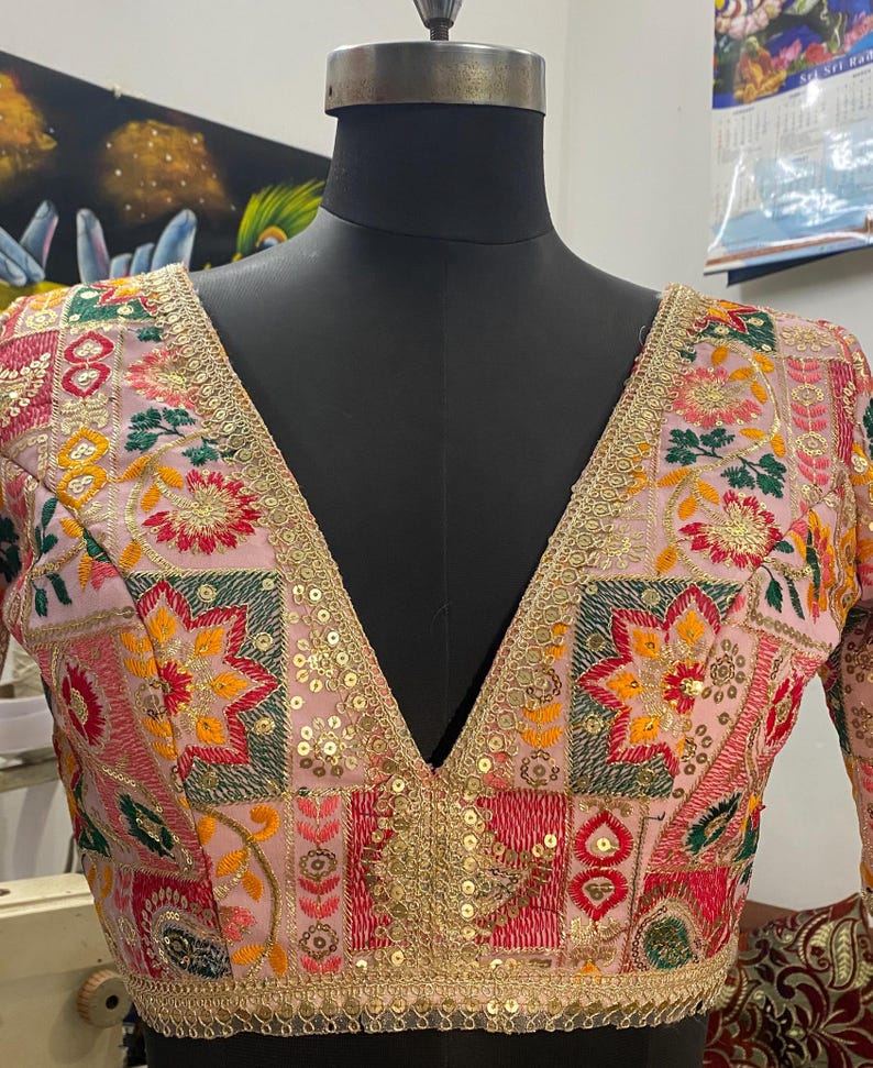 May include: A pink, embroidered blouse with a deep V-neckline. The blouse features a patchwork design with floral and geometric motifs in shades of red, green, yellow, and gold. The neckline and sleeves are trimmed with gold sequins.