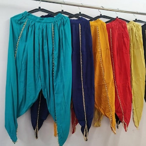 May include: A row of colorful, pleated pants hanging on black hangers. The pants come in teal, navy, yellow, red, gold, and black. Each pair has gold beaded trim along the sides and bottom.