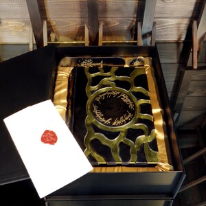 May include: A black box containing a book with gold detailing and text. A white card with a red wax seal is placed next to the book. The book is in a black box with gold satin lining.