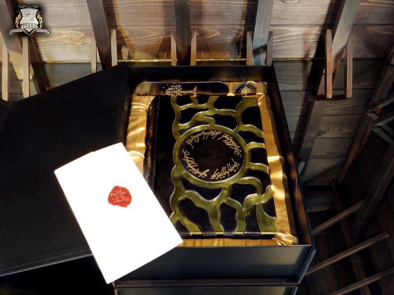 May include: A black box containing a book with gold detailing and text. A white card with a red wax seal is placed next to the book. The book is in a black box with gold satin lining.