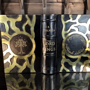 May include: A decorative book titled "The Lord of the Rings" by J.R.R. Tolkien. The book features a black cover with gold detailing and circular designs. The spine is black with gold lettering.