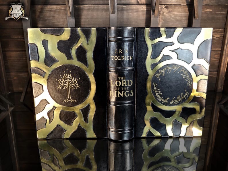 May include: A decorative book titled "The Lord of the Rings" by J.R.R. Tolkien. The book features a black cover with gold detailing and circular designs. The spine is black with gold lettering.