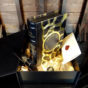 May include: A black box set featuring a book titled "The Lord of the Rings" by J.R.R. Tolkien. The book has gold accents and is accompanied by a bookmark, a key chain, and a sealed envelope.