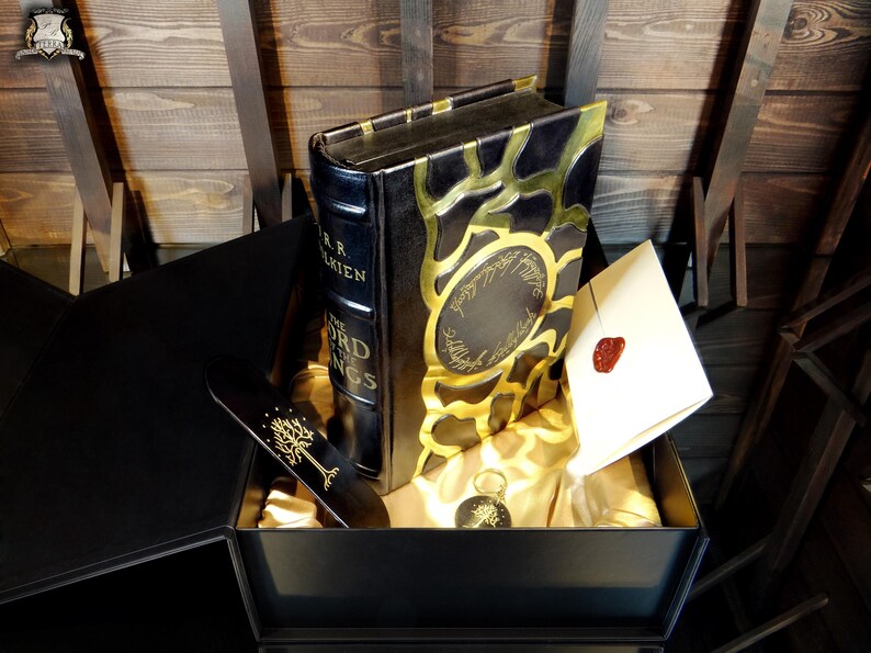 May include: A black box set featuring a book titled "The Lord of the Rings" by J.R.R. Tolkien. The book has gold accents and is accompanied by a bookmark, a key chain, and a sealed envelope.