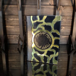 May include: A decorative book with a black cover and gold detailing. The cover has a gold circular design with text and a gold pattern. The book is set against a wooden backdrop.