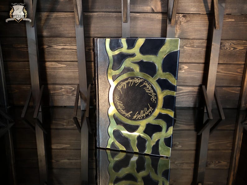 May include: A decorative book with a black cover and gold detailing. The cover has a gold circular design with text and a gold pattern. The book is set against a wooden backdrop.