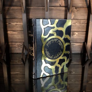 May include: A hardback book with a black cover and gold detailing. The cover has a gold sunburst design and circular text. The book's spine reads "THE LORD OF THE RINGS".