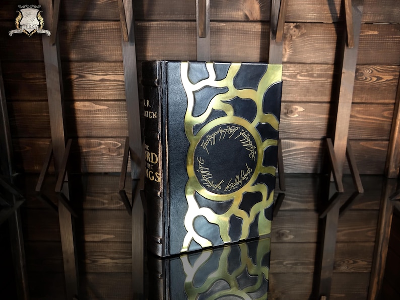 May include: A hardback book with a black cover and gold detailing. The cover has a gold sunburst design and circular text. The book's spine reads "THE LORD OF THE RINGS".