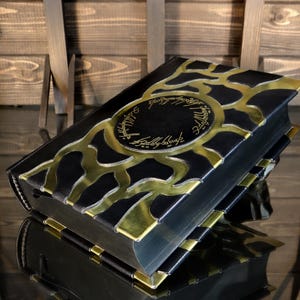 May include: A black book with gold detailing and text. The cover has a gold design and text in a circular pattern. The book is on a reflective surface, with a wooden backdrop.