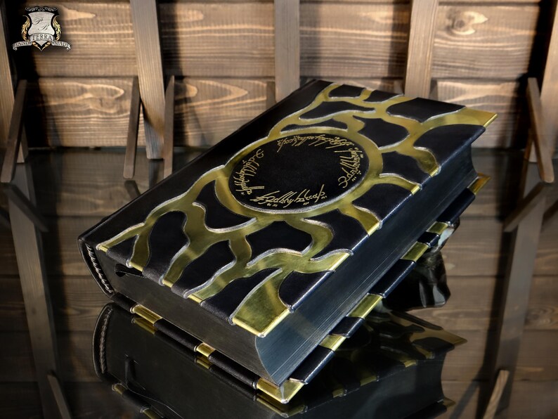 May include: A black book with gold detailing and text. The cover has a gold design and text in a circular pattern. The book is on a reflective surface, with a wooden backdrop.