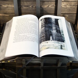 May include: An open book with a detailed illustration of a dark tower on the right page. The book is propped up on a wooden stand with a black ribbon bookmark. The pages are thick and the book has a vintage aesthetic.