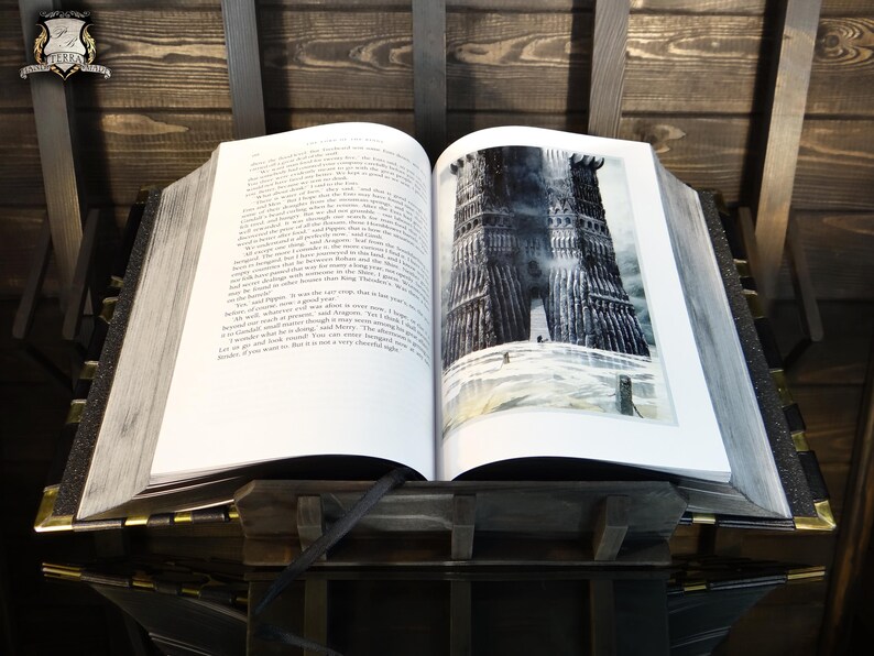 May include: An open book with a detailed illustration of a dark tower on the right page. The book is propped up on a wooden stand with a black ribbon bookmark. The pages are thick and the book has a vintage aesthetic.