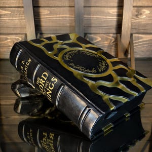 May include: A black and gold decorative box shaped like a book, with the title "The Lord of the Rings" and the author's name, J.R.R. Tolkien, visible on the spine. The box has a metallic gold design on the cover and spine.