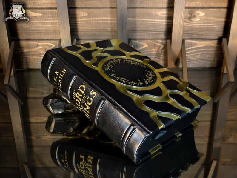 May include: A black and gold decorative box shaped like a book, with the title "The Lord of the Rings" and the author's name, J.R.R. Tolkien, visible on the spine. The box has a metallic gold design on the cover and spine.